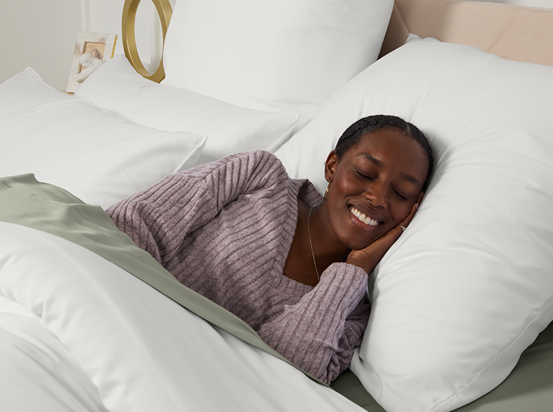 Vrouw slaapt vredig in bed met witte en saliegroene lakens, draagt een lavendelkleurige trui en glimlacht ontspannen.