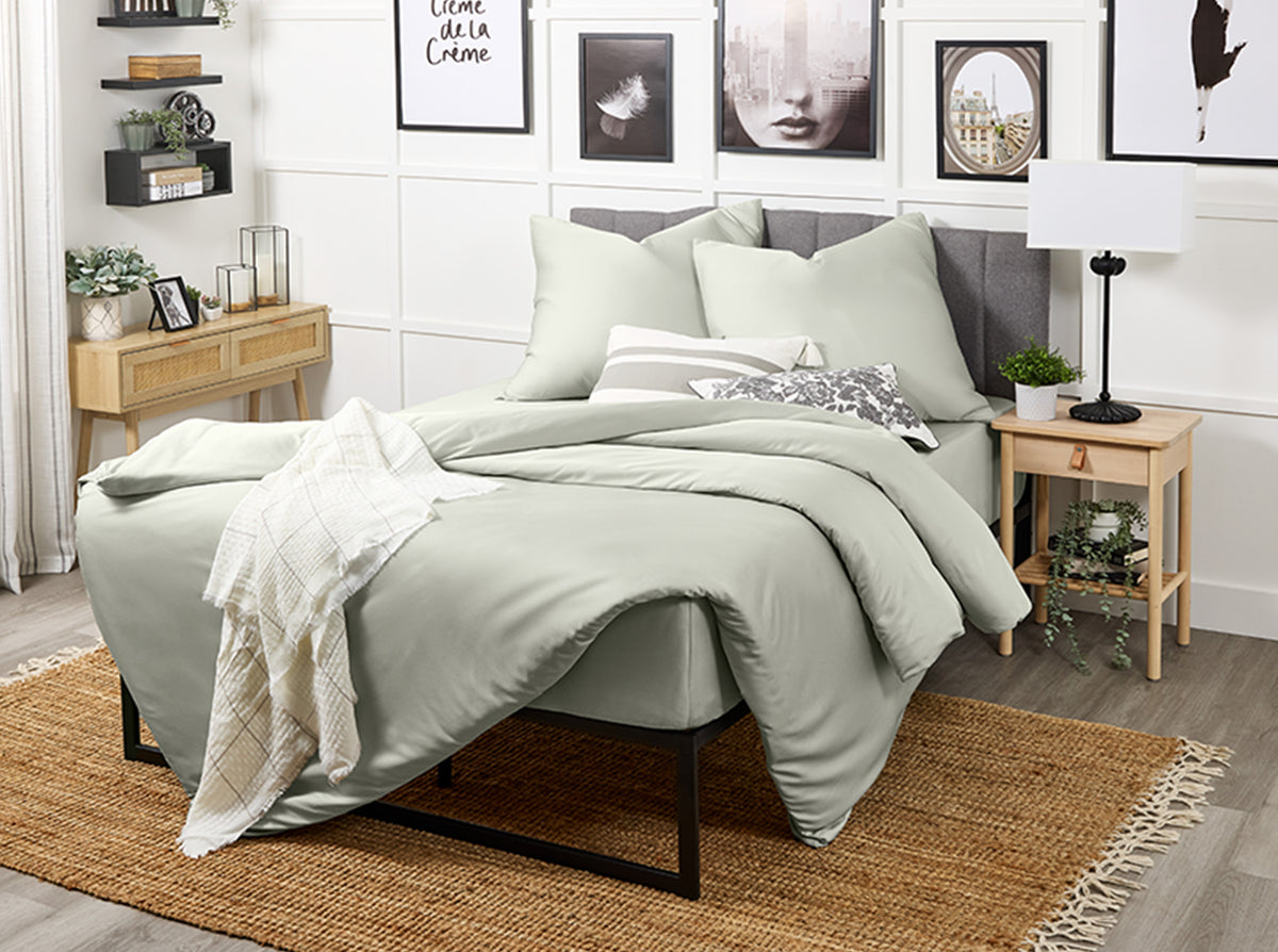 Netjes opgemaakt bed met saliegroen beddengoed in een moderne slaapkamer met muurdecoratie en planten.