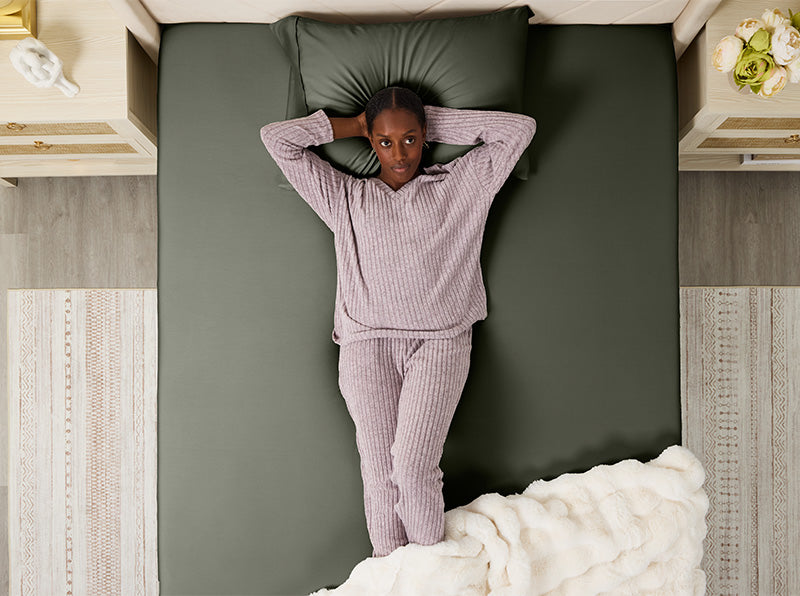 Vrouw ligt op een saliegroen hoeslaken, handen achter het hoofd, in comfortabele lavendelkleurige pyjama op een netjes opgemaakt bed.