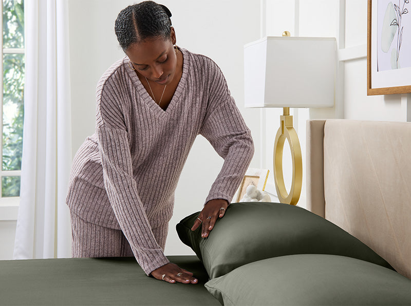 Vrouw in lavendelkleurige loungewear strijkt saliegroene lakens glad op een netjes opgemaakt bed in een lichte, moderne slaapkamer.